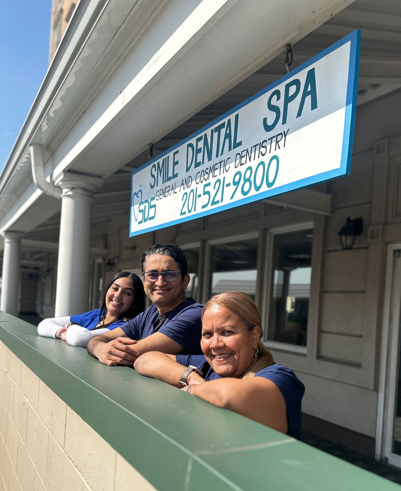 The image shows three individuals leaning against a railing in front of a building with a sign that reads Smile Dental Spa.